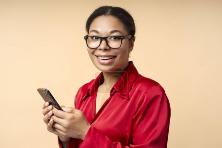 Téléchargez les photos : Portrait de Afro-Américain souriant portant des lunettes tenant téléphone portable faisant des achats en ligne isolé sur fond beige, espace de copie. Femme d'affaires prospère utilisant smartphone, services bancaires mobiles - en image libre de droit