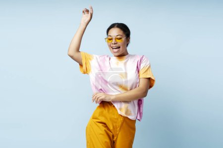 Foto de Mujer afroamericana feliz bailando con camiseta de moda y gafas aisladas sobre fondo azul. Sonriente modelo de moda elegante que se divierten posando para la imagen en el estudio. Concepto de verano - Imagen libre de derechos