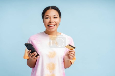 Téléchargez les photos : Portrait de jeune femme afro-américaine souriante tenant une carte de crédit, en utilisant l'application mobile shopping en ligne isolé sur fond bleu. Joyeuse femme élégante commande de nourriture. Concept technologique - en image libre de droit