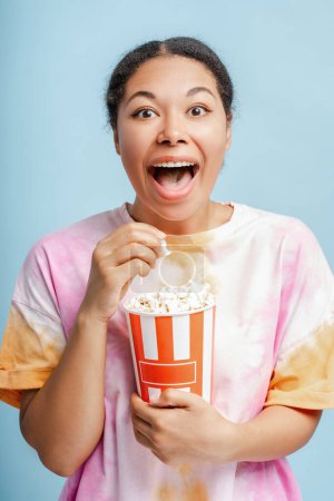Photo for Excited African American woman holding bucket with popcorn, eating snacks watching movie with open mouth in isolated on blue background. Cinema time, advertisement concept - Royalty Free Image