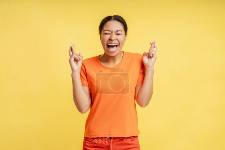 Photo for Beautiful stylish stunning African American woman wearing orange casual shirt, closing her eyes and fingers crossed, expressing positivity and hopefulness, isolated over yellow background - Royalty Free Image