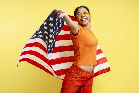 Foto de Hermosa mujer afroamericana sonriente sosteniendo la bandera americana aislada sobre fondo amarillo. Retrato de la celebración patriota feliz joven Día de la Independencia mirando hacia otro lado. 4 de julio concepto - Imagen libre de derechos