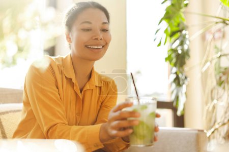 Téléchargez les photos : Portrait d'une belle femme afro-américaine souriante tenant une limonade fraîche savoureuse ou un cocktail assis et relaxant dans une caffe moderne. Concept d'été - en image libre de droit