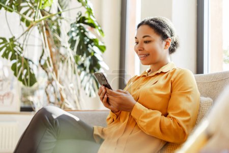 Foto de Feliz sonriente mujer de negocios afroamericana, freelancer sosteniendo teléfono móvil, trabajando en línea desde casa. Elegante redactor con teléfono inteligente sentado en la cafetería. Negocios exitosos - Imagen libre de derechos