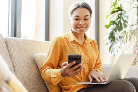 Téléchargez les photos : Femme d'affaires afro-américaine souriante moderne, pigiste tenant un téléphone mobile, utilisant un ordinateur portable fonctionnant en ligne à partir de la maison. Élégant rédacteur avec smartphone assis dans un café. Une entreprise prospère - en image libre de droit