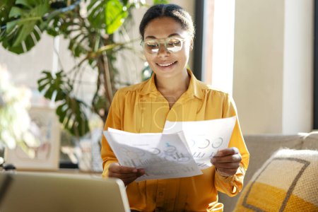 Photo for Beautiful smiling African American business woman, freelancer wearing stylish eyeglasses holding documents using laptop working online from home. Successful business - Royalty Free Image