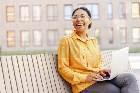 Photo for Confident smiling African American businesswoman using laptop working online looking away sitting on bench. Freelancer copywriter typing on keyboard planning project. Technology, successful business - Royalty Free Image