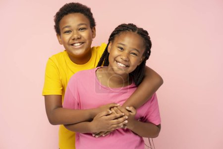 Foto de Retrato de un niño y una niña afroamericanos sonrientes con una camiseta colorida abrazándose aislados sobre un fondo rosa - Imagen libre de derechos