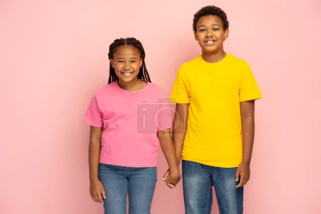 Photo for Young smiling African American kids, brother and sister wearing pink and yellow t shirts holding hands standing together isolated on pink background. Friendship concept - Royalty Free Image