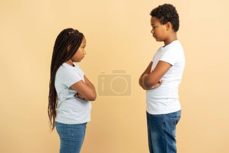 Photo for Portrait of offended children African American brother and sister standing and looking at each other with crossed arms isolated on beige background, copy space. Family concept - Royalty Free Image