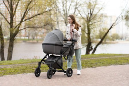 Photo for Positive young woman pushing stroller with baby in park in city. Beautiful mother taking care of her child, outdoor walking, parenthood - Royalty Free Image