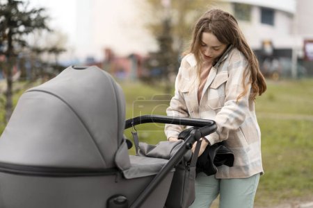 Photo for Smiling woman talking on mobile phone, pushing pram with baby in park. Young mother caring about health, communication, holding smartphone - Royalty Free Image