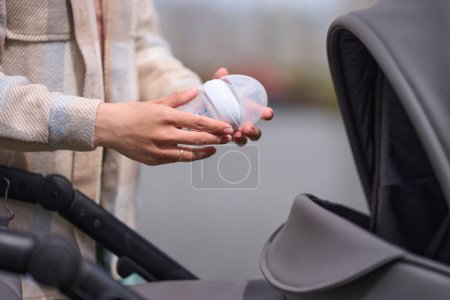 Photo for Selective focus on a baby bottle with milk in the hands of a young mother over a stylish gray stroller. Close-up female hands holding infant formula bottle above a baby carriage on nature background. - Royalty Free Image