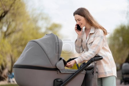 Photo for Smiling, cheerful mother talking on mobile phone, looking into stroller at newborn baby. Attractive woman walking outdoors, motherhood, parenthood, child care - Royalty Free Image