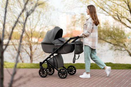 Photo for Young woman pushing stroller, walking outdoors in park. Smiling, happy mother with baby in carriage - Royalty Free Image