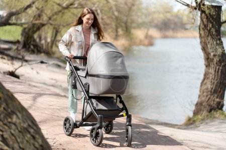 Foto de Moderna, joven madre caminando empujando cochecito con bebé al aire libre, caminando en el parque. Mujer feliz, día soleado, maternidad alegre - Imagen libre de derechos