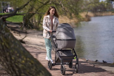 Photo for Young woman, loving mother pushing gray baby stroller and slowly walking at town green park near a lake, in warm sunny spring day. Spending time with infant and breathing fresh air. Enjoying stroll. - Royalty Free Image