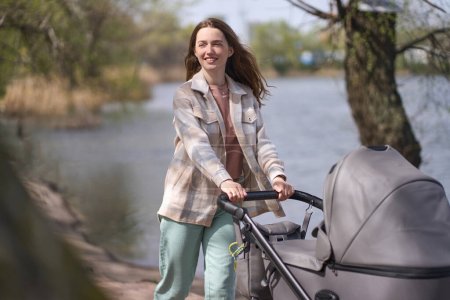 Photo for Charming Caucasian young woman, mom walking in the park by lake with her newborn baby in buggy, enjoying breathing fresh air in the nature on a beautiful warm sunny day. Maternity. Healthy lifestyle - Royalty Free Image