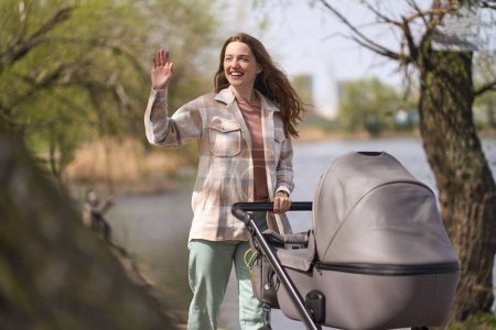 Photo for Caucasian red haired beautiful young woman, loving caring mother waving hello with her hand, smiles looking away, enjoy stroll with her newborn baby in stylish gray buggy, in the sunny park by lake - Royalty Free Image