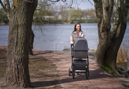 Photo for Portrait of attractive cheerful young red haired Caucasian woman, loving caring happy mother walking newborn carriage, resting on fresh air, spending free time in the nature by lake on warm sunny day - Royalty Free Image