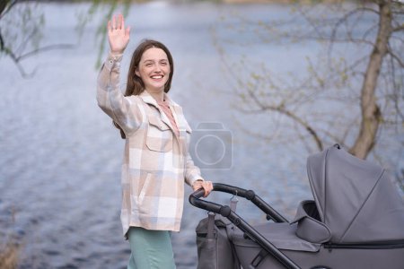 Photo for Loving caring mother waving hello with hand, smiles looking away, walking with her newborn child in stylish gray eco leather stroller in the park by river, on a beautiful warm sunny spring day - Royalty Free Image
