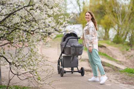 Photo for Young woman - mother smiles at camera while pushes baby stroller, walks at green park, breathing fresh air, enjoying stroll on warm sunny spring day. Leisure activity. Healthy lifestyle. Maternity - Royalty Free Image