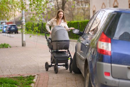 Photo for Young mother pushing stroller with her newborn child, feels outraged by an incorrectly parked car on a pedestrian sidewalk, blocking the passage of pedestrians walking down the street, with baby prams - Royalty Free Image