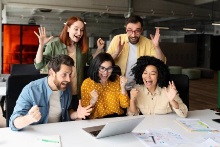 Happy team of diverse business people clenching fists from excitement, winning tender, goals achievement, rejoicing sitting at desk with laptop, graphs and diagrams in modern corporate office building