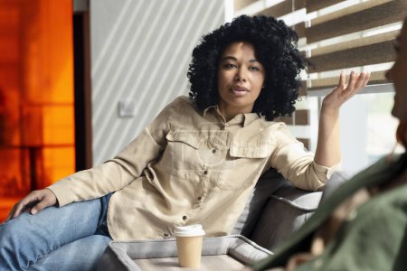 Téléchargez les photos : Portrait d'une femme afro-américaine détendue vêtue de vêtements décontractés, assise sur le canapé, parlant avec sa collègue tout en profitant de sa pause café dans un intérieur moderne de bureau d'affaires - en image libre de droit