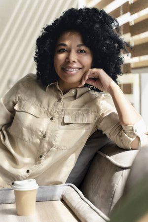 Foto de Auténtico retrato de una joven afroamericana positiva, trabajadora de oficina, gerente de ventas, sonriendo mirando a la cámara, disfrutando de su pausa para tomar café en el interior de la oficina moderna. Personas, carrera y estilo de vida - Imagen libre de derechos
