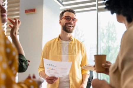 Téléchargez les photos : Bel homme barbu caucasien dans des lunettes élégantes, tenant document avec des diagrammes de croissance des ventes, souriant parler à ses collègues, partager des idées et des stratégies de développement des affaires dans le bureau moderne - en image libre de droit