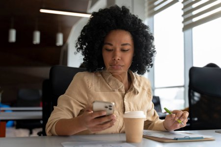 Téléchargez les photos : Jeune femme afro-américaine confiante, gestionnaire de bureau utilisant le téléphone mobile, communication en ligne avec des partenaires d'affaires, assis sur le lieu de travail avec une tasse de café chaud jetable dans l'intérieur du bureau moderne - en image libre de droit