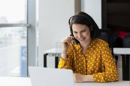 Pleasant young businesswoman, sales manager, executive director or recruiter in audio headset, sitting at table, using laptop having online meeting with partners or job interviews in office interior