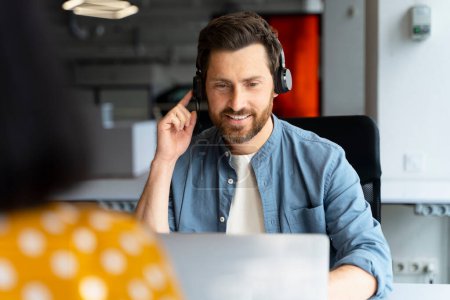 Hombre barbudo positivo con propósito atractivo seguro, usando auriculares, gerente de centro de llamadas hablando con clientes o clientes a través de videoconferencia en línea, sentado en el escritorio con computadora portátil en el interior de la oficina