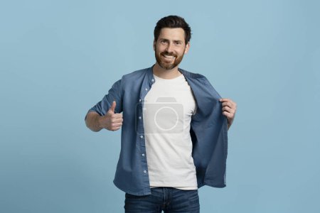 Photo for Handsome middle aged Caucasian bearded man in casual denim and white mockup t-shirt, gesturing with thumb up looking at camera, isolated over blue color background - Royalty Free Image
