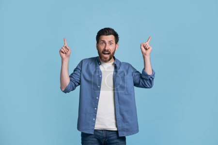 Photo for Amazed middle aged Caucasian bearded man 40s, in casual denim clothes and t-shirt, pointing his index fingers up, expressing WOW emotion, amazement and surprise, isolated on blue color background - Royalty Free Image