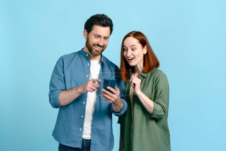 Kaukasisch lächelndes erwachsenes Paar, das sein Mobiltelefon benutzt, Nachrichten scrollt, Inhalte sozialer Medien überprüft, Online-Nachrichten versendet und neue mobile Anwendungen überprüft. Isolierte blaue Farbe Studio Hintergrund