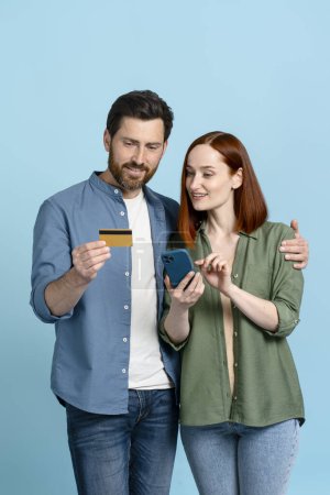 Kaukasisches glückliches lächelndes Paar mit Plastikkreditkarte und Handy, isoliert auf blauem Hintergrund. Online-Shopping. Internet-Banking. Bargeldloses Bezahlen. Rechnungen bezahlen. Buchung und Essensbestellung