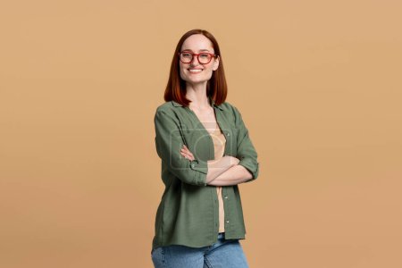 Téléchargez les photos : Portrait studio d'une rousse confiante aux cheveux raides, portant des lunettes tendance, souriante regardant la caméra, posant les bras croisés, isolée sur fond beige. Concept de personnes - en image libre de droit