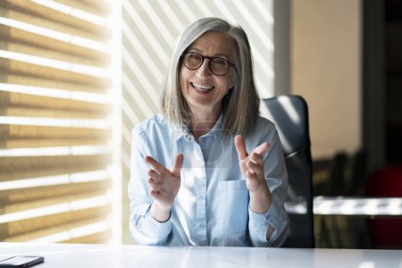 Photo for Caucasian gray haired elegant mature woman gestures with her hands, smiles and talks looking toward the camera, sitting at desk in modern office interior, with shadows of the window blinds on her face - Royalty Free Image