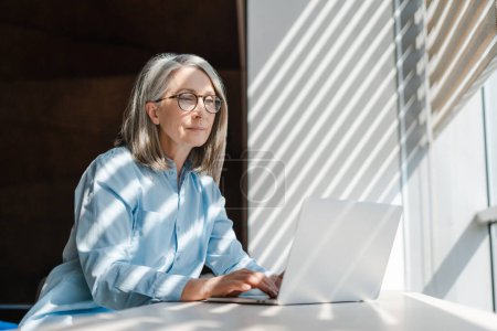 Téléchargez les photos : Pensive femme d'affaires senior utilisant ordinateur portable travaillant en ligne assis dans un bureau moderne - en image libre de droit