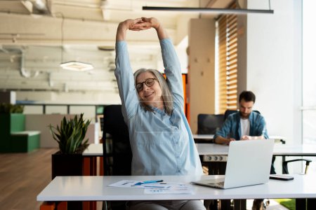 Attractive smiling senior woman stretching arms and back, relaxing, taking break working in modern office. Successful confident businesswoman finish work 