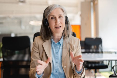 Caucasian elegant gray haired mature adult woman, call center manager wearing audio headset headphones, smiling and talking looking at camera. Ideal mockup for insert in laptops and digital screens