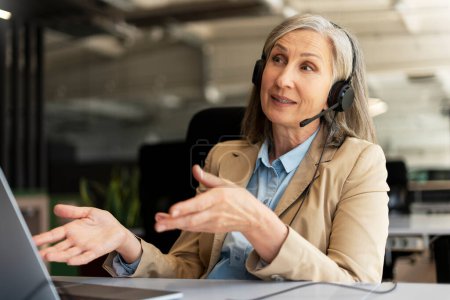 Confident gray haired senior businesswoman, CEO, wearing headphones, talking to clients, investors or customers via online video conference, sitting at desk with laptop in office modern interior
