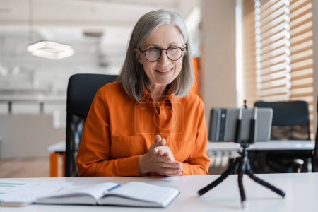Confident smiling senior woman using mobile phone communication online, having video call. Successful gray haired influencer recording video, looking at smartphone display, sitting at workplace 
