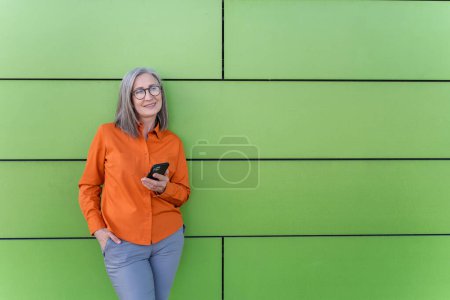 Photo for Portrait smiling senior woman holding smartphone using mobile app shopping online standing on the street looking away, copy space. Mobile banking technology concept - Royalty Free Image