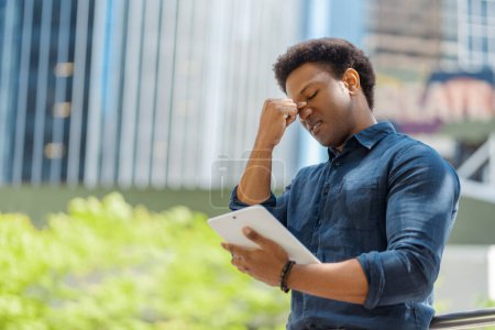 Foto de Hombre de negocios afroamericano cansado sosteniendo tableta digital, comprobar el correo electrónico, trabajar en línea, leer noticias de pie en la calle. Estudiante serio estudiando, aprendiendo el idioma en el campus universitario - Imagen libre de derechos