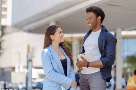 Photo for African male and latin female colleagues standing at street and discussing, smiling. Business partners talking outdoors - Royalty Free Image
