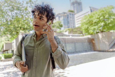 Photo for Portrait of smiling handsome hispanic student talking on mobile phone, answering call looking away on urban street. Successful business - Royalty Free Image