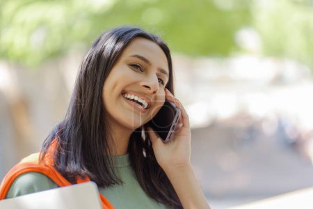 Foto de Primer plano sonriendo estudiante indio hablando teléfono móvil, contestando llamada mirando hacia otro lado en la calle de la ciudad. Sonriendo joven mujer hablando al aire libre en el espacio de copia. Educación exitosa - Imagen libre de derechos
